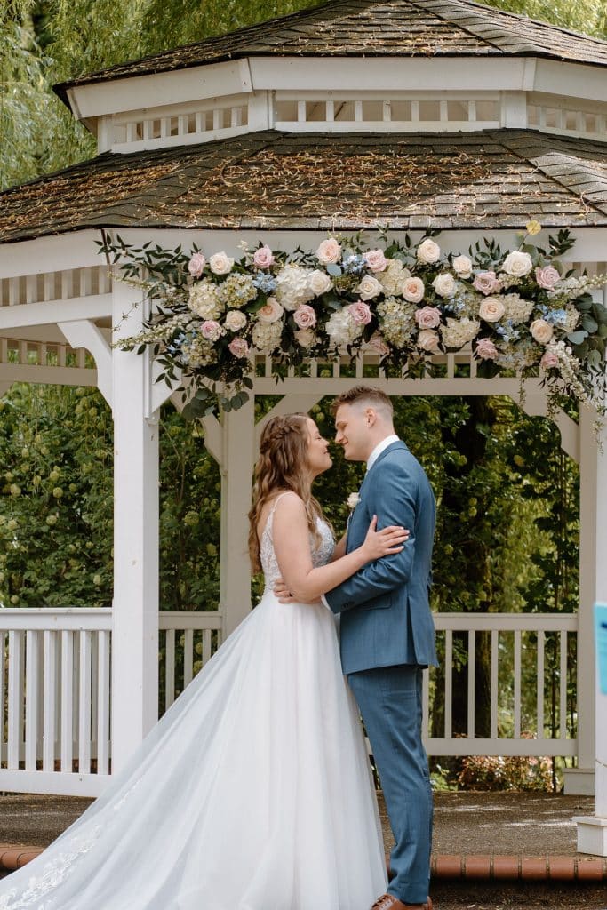 Couple embrace near a garden gazebo decorated with pastel romantic wedding flowers at Abernethy Center in Oregon City, Oregon